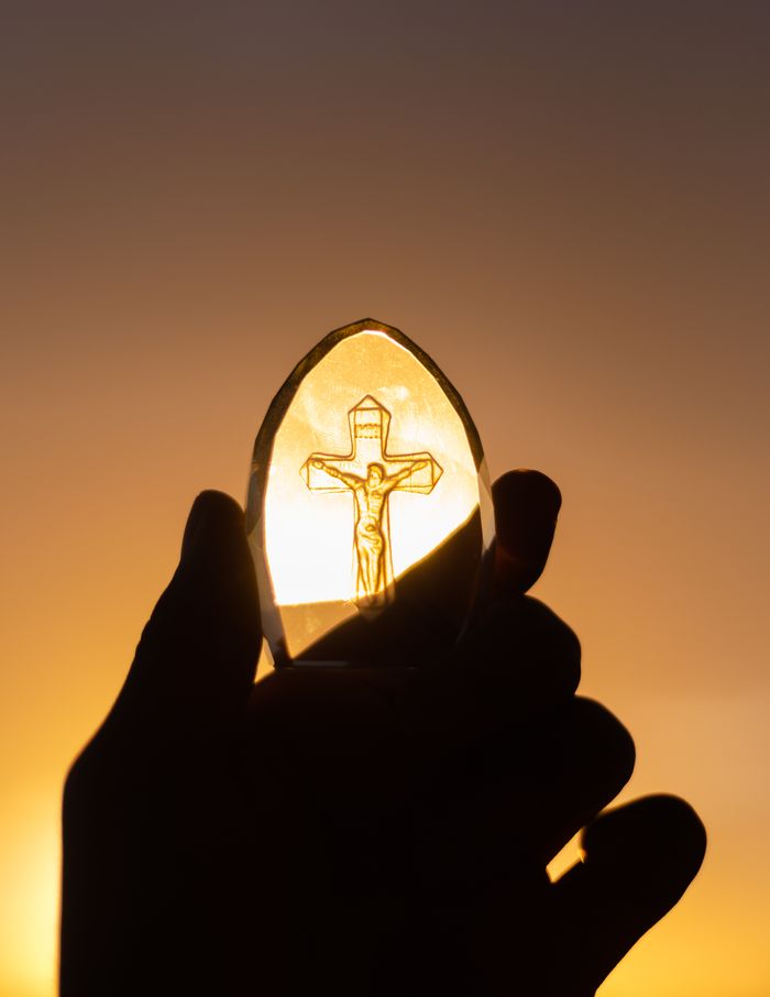 Home A silhouetted hand holds a crucifix against a vibrant sunset sky, symbolizing faith and spirituality.